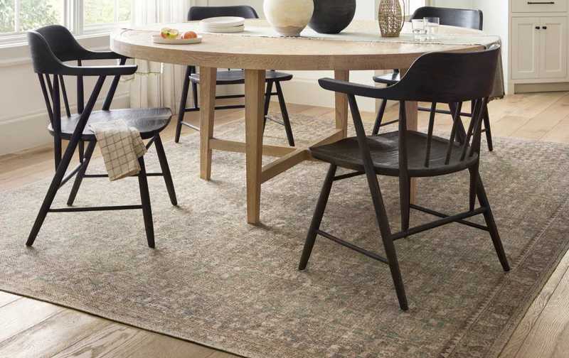 A vintage-inspired Angela Rose x Loloi Aubrey area rug with a distressed pattern and warm ombre palette anchors a modern dining set with dark chairs and a round wood table.