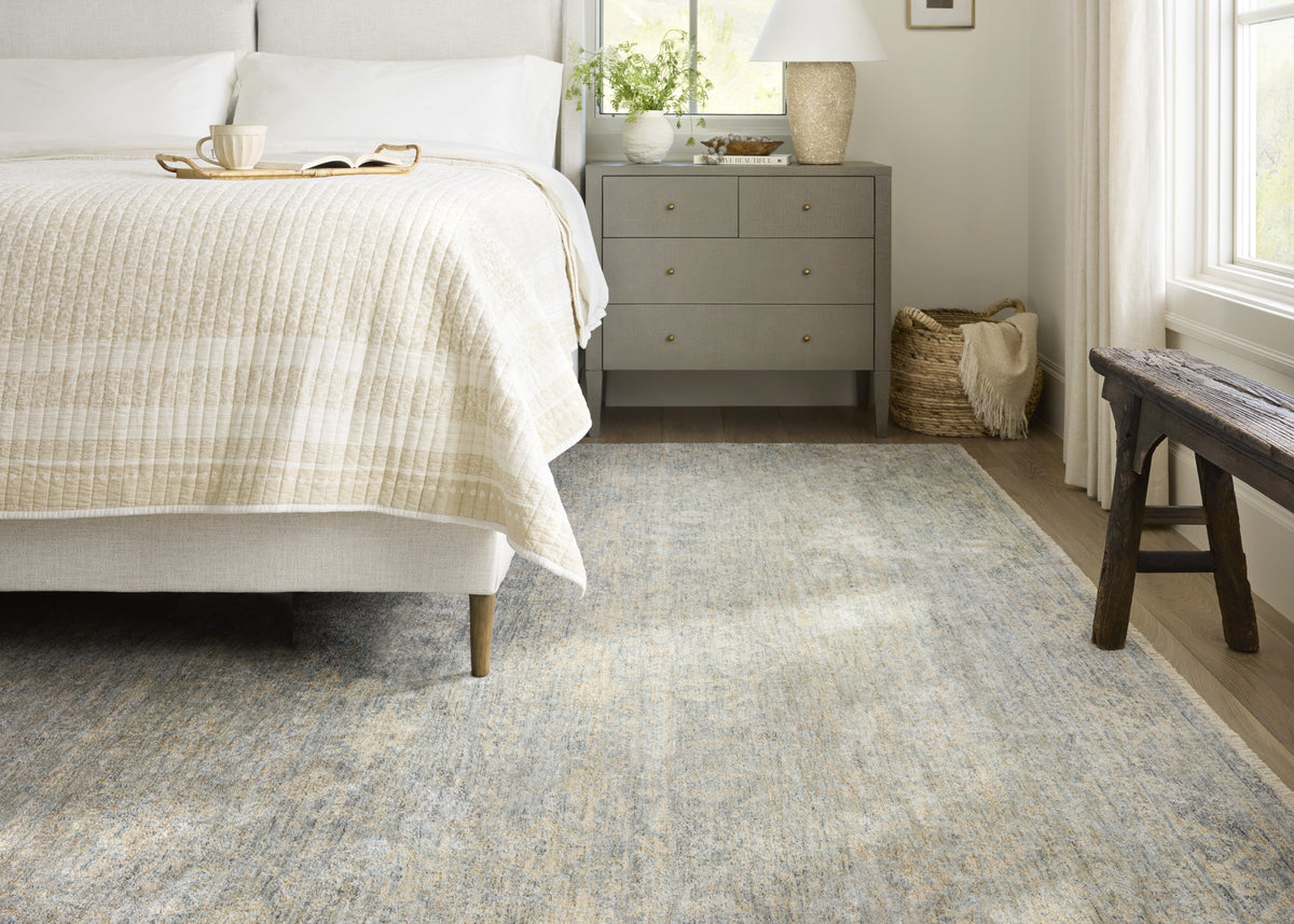 Angela Rose Blake rug by Loloi features a distressed antique design in soft blue and beige tones, placed beneath a bed in a bright bedroom.