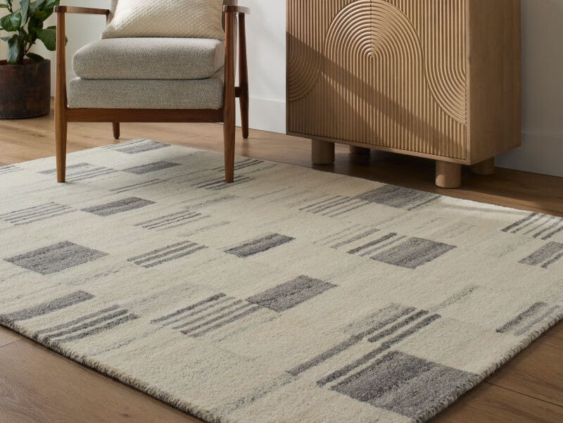 Becki Owens Max rug with grey geometric blocks on cream wool, styled under a mid-century armchair next to a slatted wood cabinet.