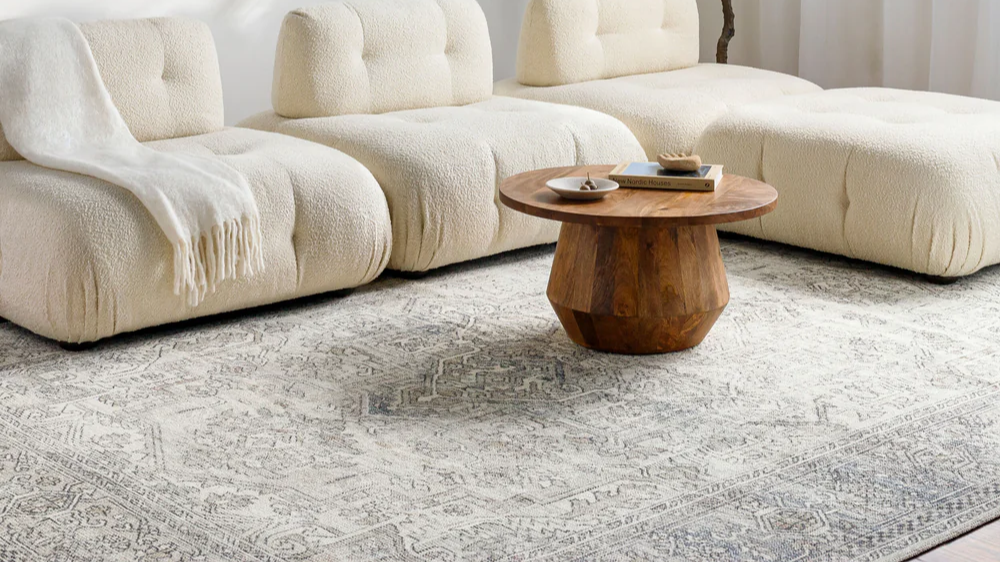Becki Owens x Livabliss Margot area rug collection in updated traditional style, featuring intricate gray patterns under a wooden side table and cream tufted sofa.