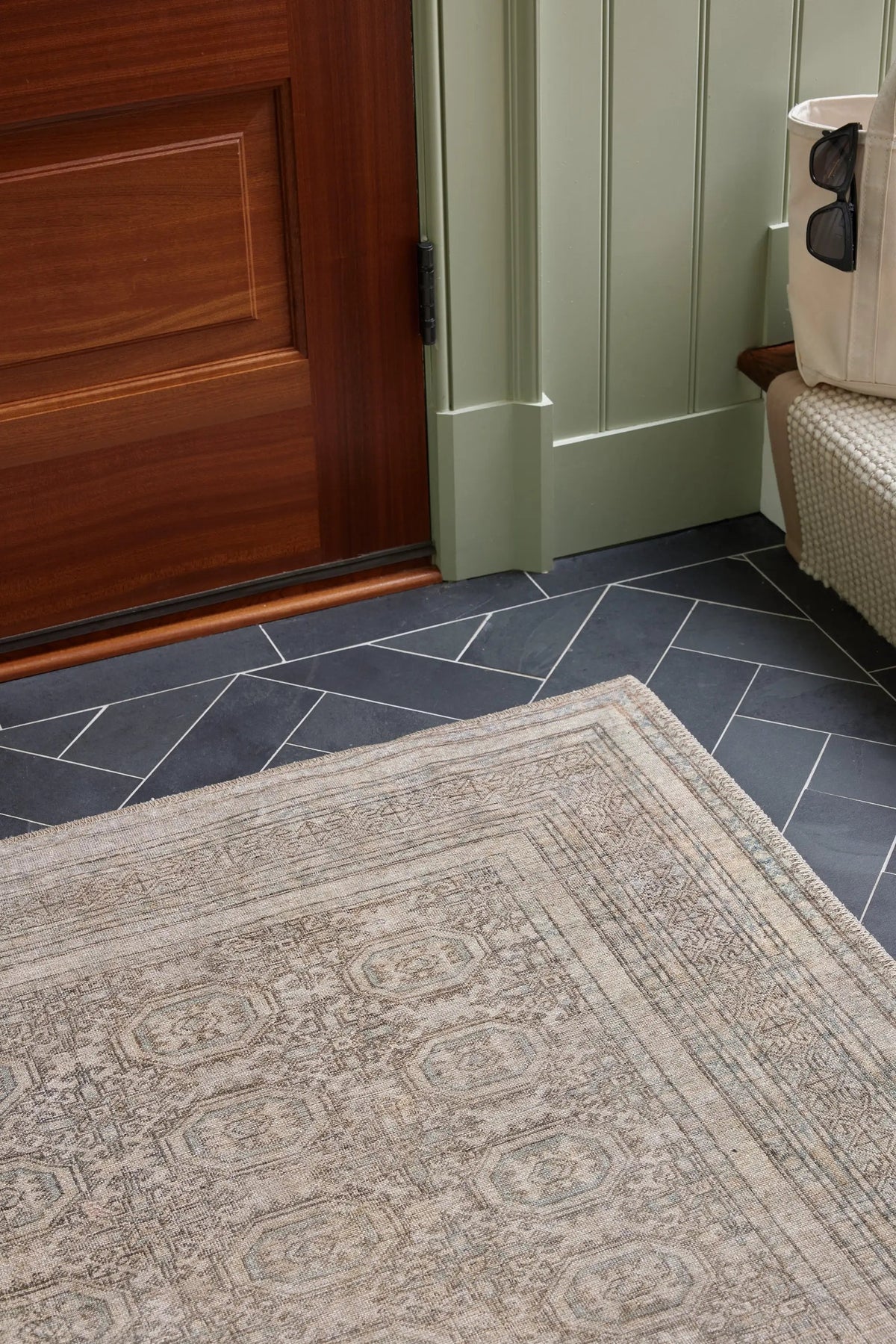 Erin Gates by Momeni Dorset DOR-1 Vintage-style green area rug with intricate borders and fringe, placed on dark slate tile near a wooden door and sage green wall.