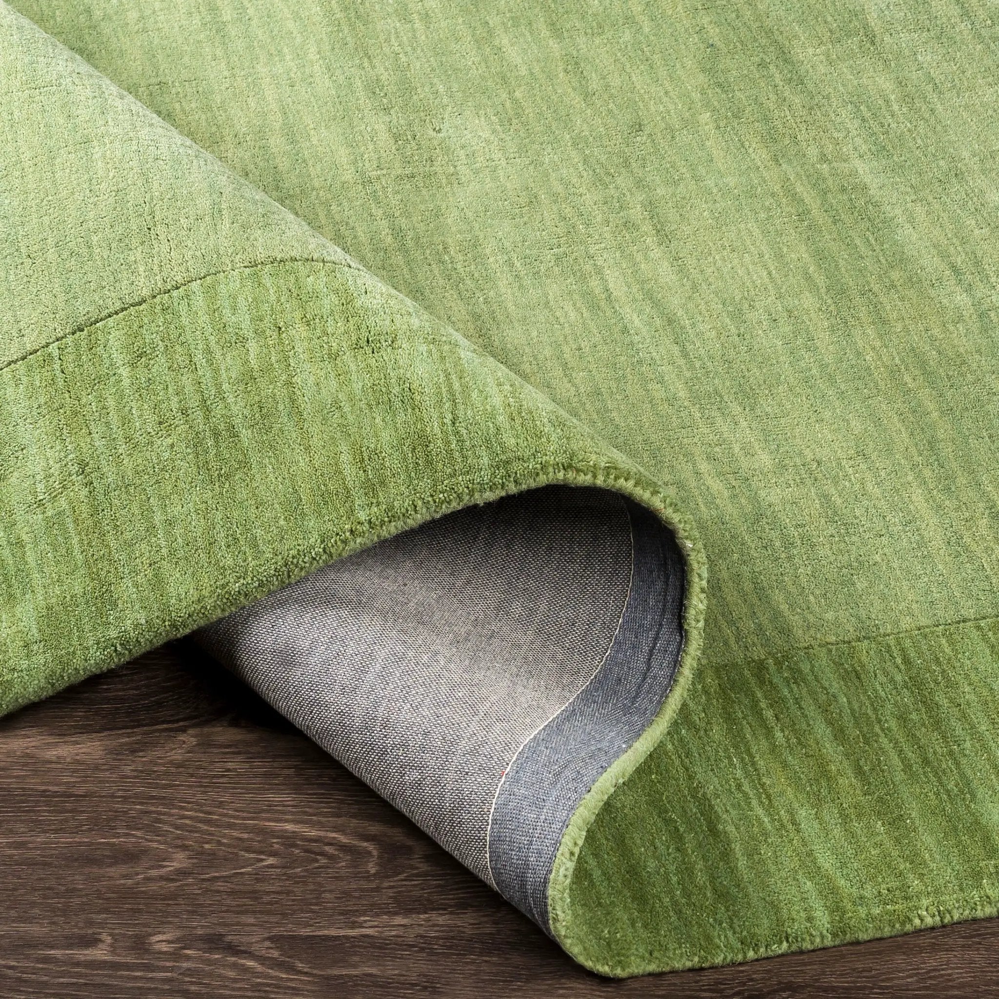 Livabliss Mystique M-310 medium green hand-loomed wool area rug with a balanced pile texture, with a curled corner showing the grey backing on a dark wood floor.