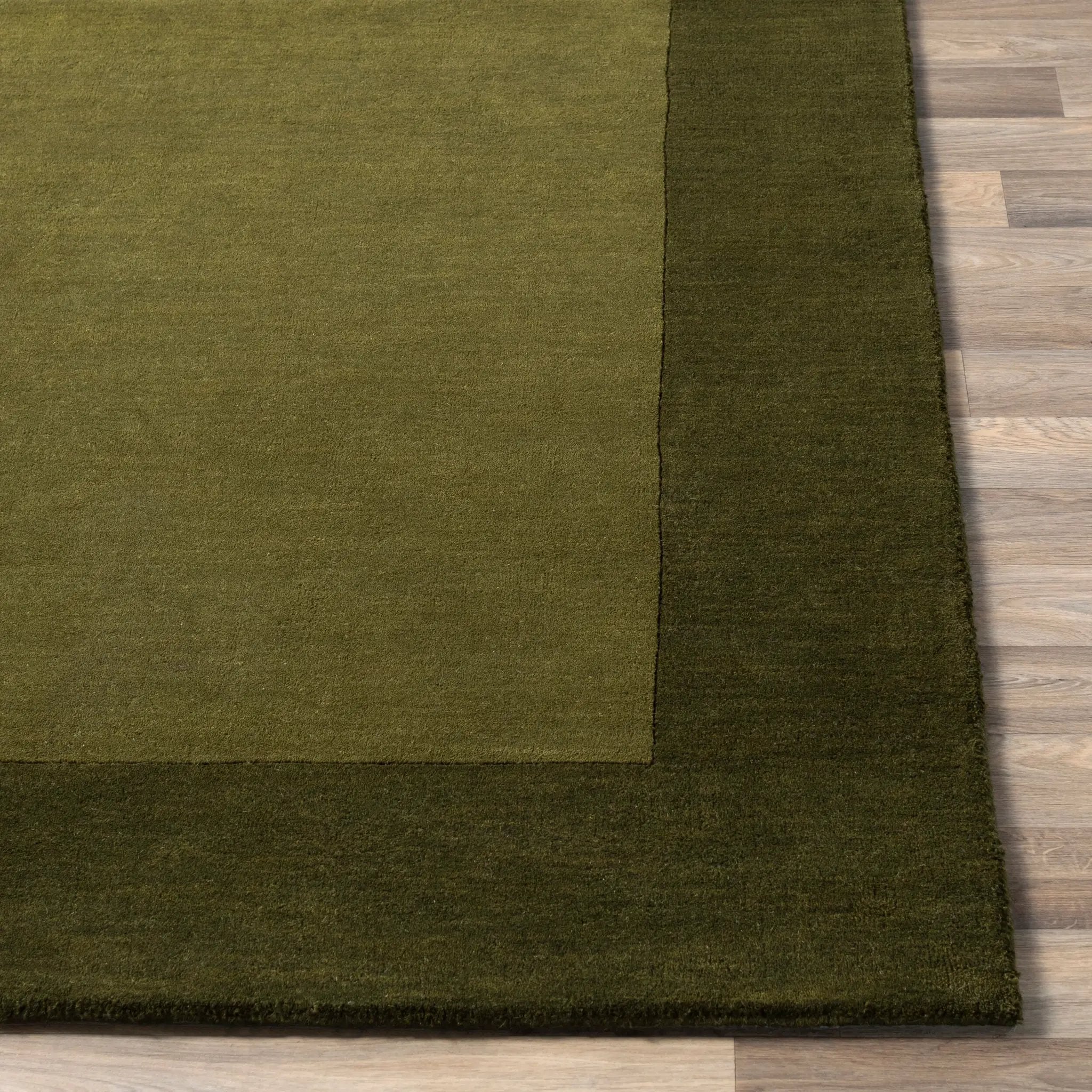 Livabliss Mystique M-315 Olive wool area rug with lighter center panel and dark border, featuring dynamic high-low pile texture on wood flooring.