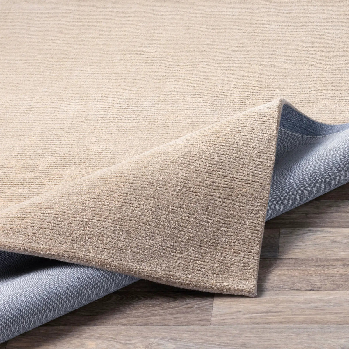 Livabliss Mystique M-335 Tan area rug with hand-loomed Indian wool construction and high-low pile texture, corner folded to reveal grey backing on wood floor.