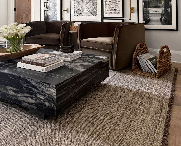 Textured beige Bohemian rug from the Galey Alix x Livabliss Bronson rug collection anchors a modern living room featuring a dark marble coffee table, brown armchairs, and a woven magazine holder.