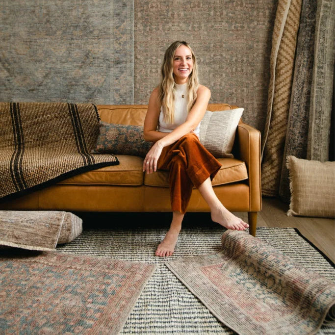 Angela Rose x Loloi area rug collection featuring designer Angela Rose seated on a caramel leather sofa, surrounded by vintage-inspired rugs in muted blue, taupe, beige, rust, and brown. Explore timeless patterns, rich textures, and warm, earthy tones to bring character and inviting style to your home at Rug-Resources.com.