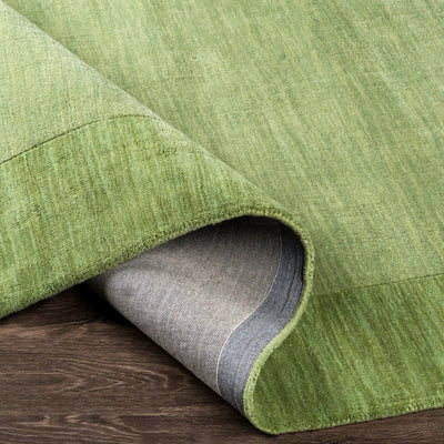 Livabliss Mystique M-310 medium green hand-loomed wool area rug with a balanced pile texture, with a curled corner showing the grey backing on a dark wood floor.