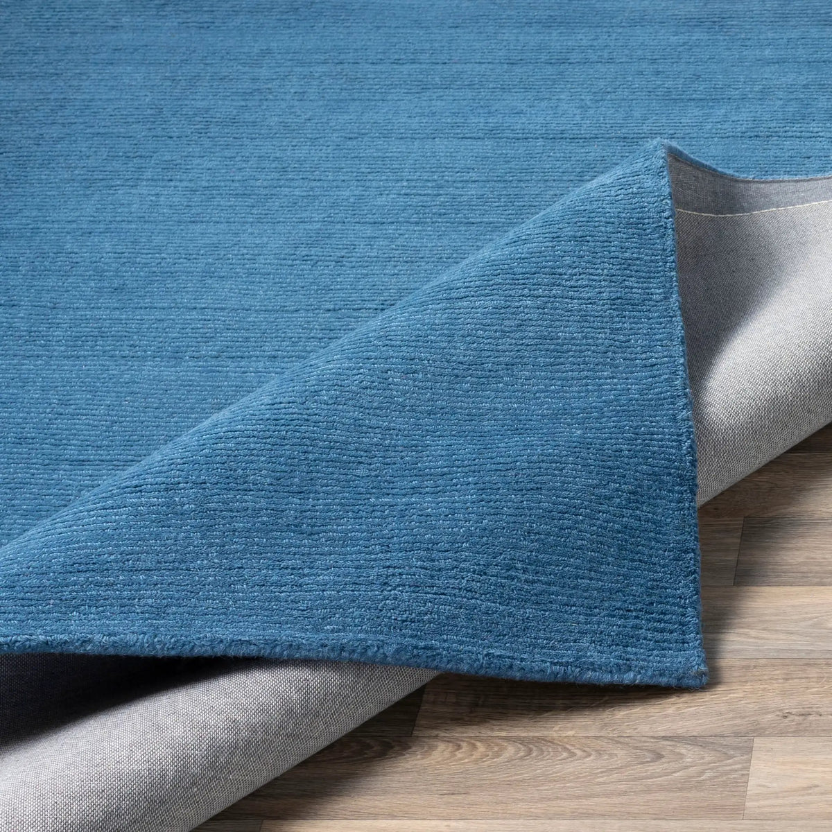 Livabliss Mystique M-330 Dark blue hand-loomed wool area rug with high-pile texture and grey backing, corner lifted to reveal construction on wood floor.