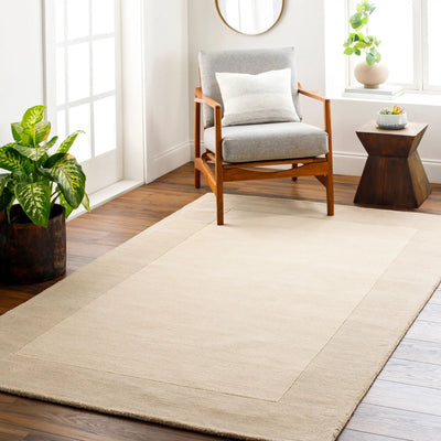 Livabliss Mystique M-348 Cream hand-loomed wool area rug with a high-low pile border, styled under a wooden armchair in a bright contemporary living space.