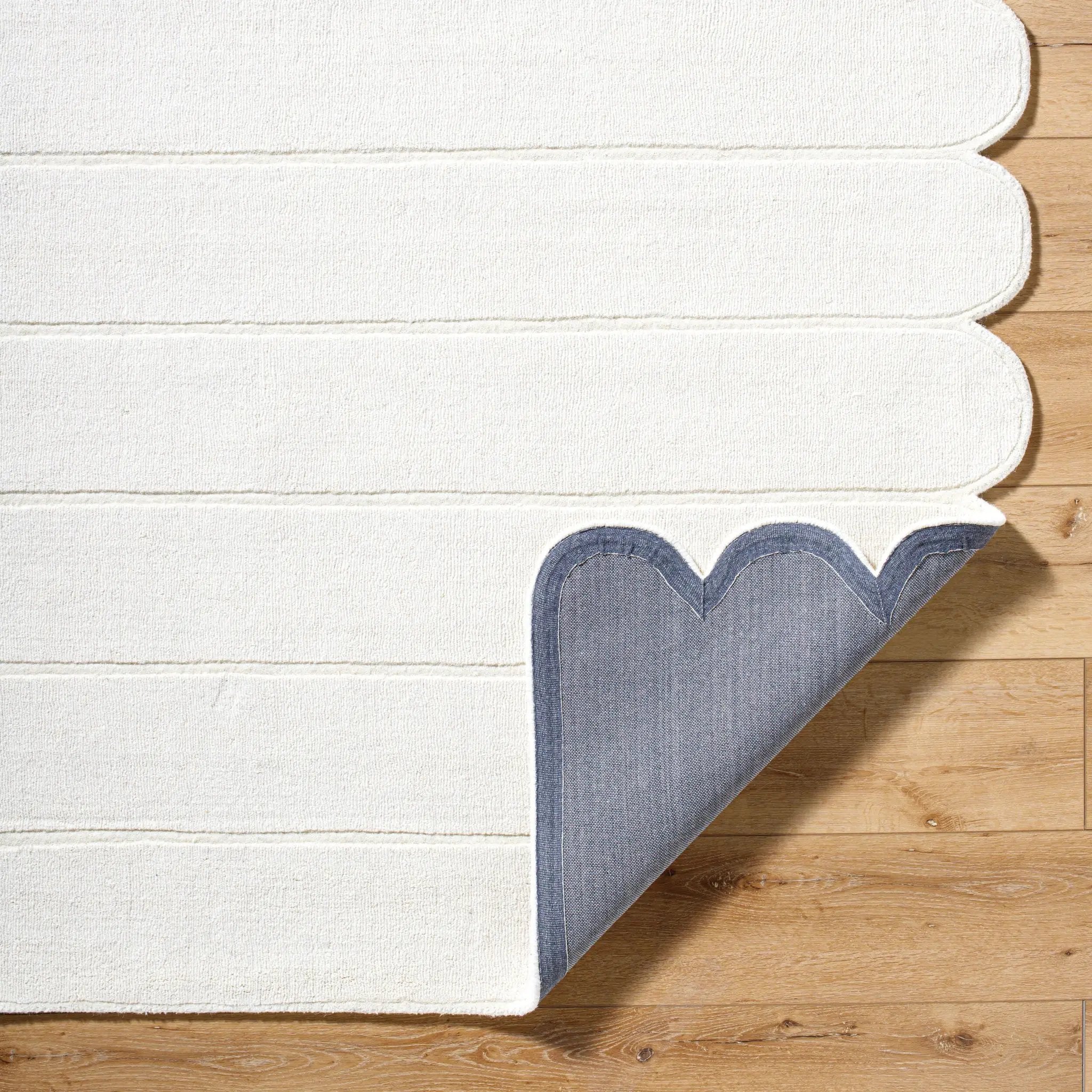 Livabliss Mystique M-5492 Ivory hand-loomed wool area rug with striped texture and scalloped edge, corner lifted to reveal blue backing on wood floor.